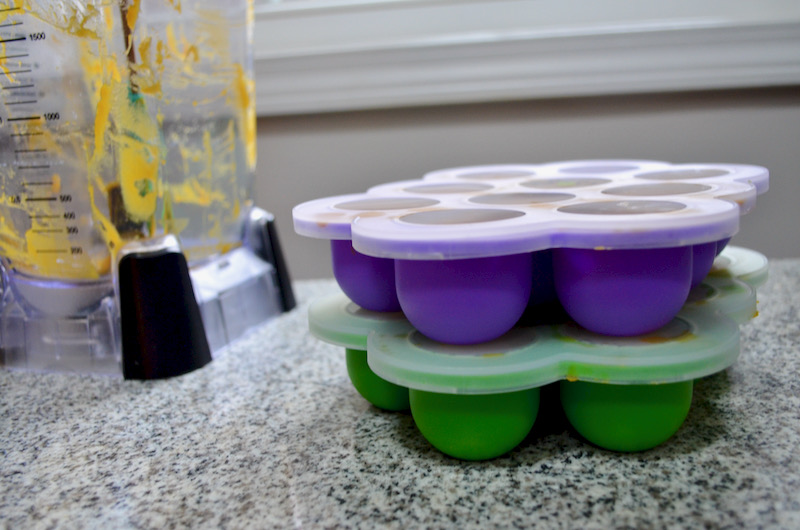 Side view of butternut squash baby food in purple and green baby food trays stacked with lids on, sitting on marble counter with blender in background