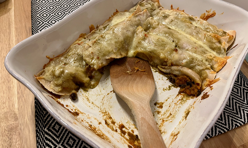 Black Bean Sweet Potato Enchiladas baked in dish with half missing and wooden spatula on wood table with placemat