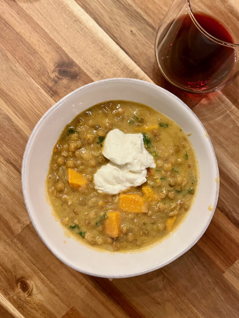 Moroccan Sweet Potato Lentil Soup in white bowl served with a dollop of greek yogurt and a glass of red wine sitting on wood table