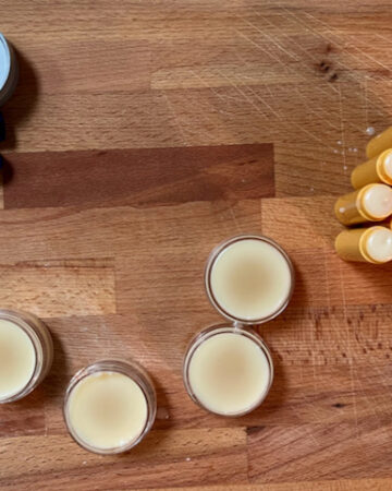 sitting on a wood cutting board, lip balm recipe poured into tubes and tubs in the drying stage, it is more cream in color