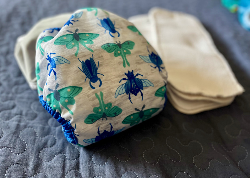 Cloth diaper with bug print displayed on blue background next two white cloth diaper inserts