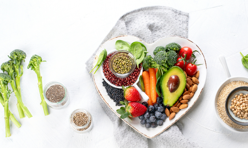 A vegetarian plate. The center of image is white heart shaped plate with lost of colorful veggies, fruit, and grains. The sides of the image have extra broccoli, seeds, and grains. 